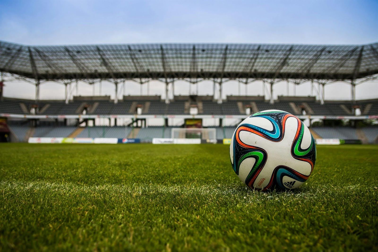 A colorful soccer ball rests on a grass field, with empty stadium seating in the background. 